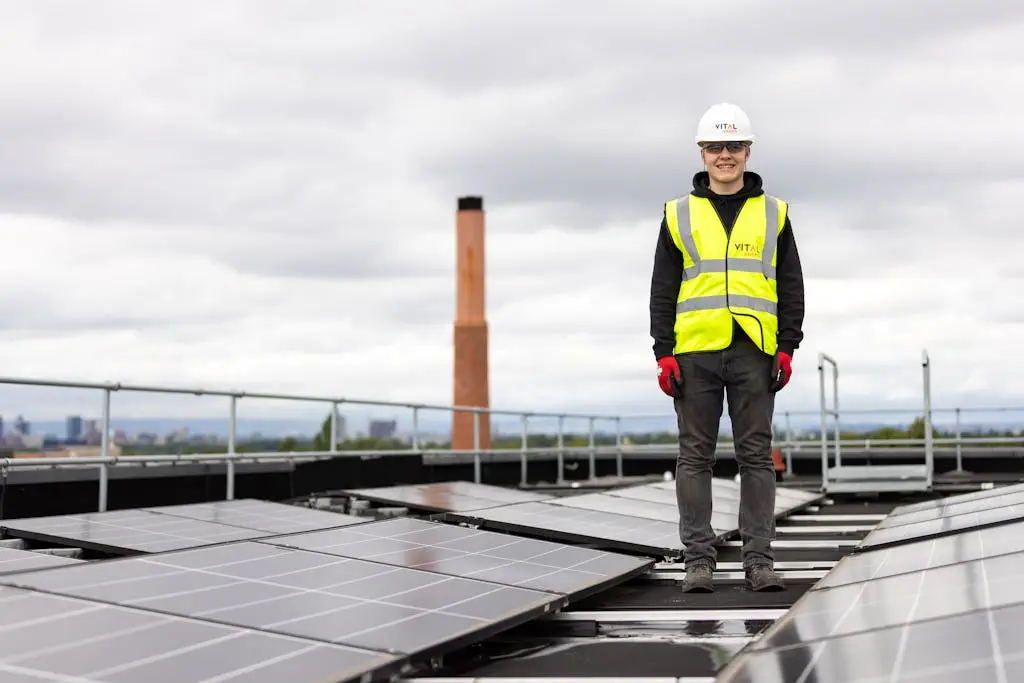 an engineer in a hard hat and safety vest inspects solar panels on a rooftop. 19895867 An engineer in a hard hat and safety vest inspects solar panels on a rooftop.
